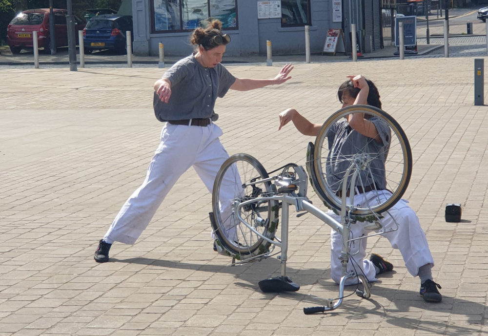Two dancers stare at an upturned bicycle