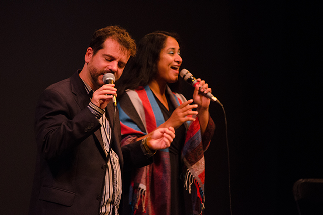 A man and a woman sing into microphones on stage