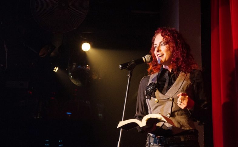 A performer with red hair speaks into a microphone on stage